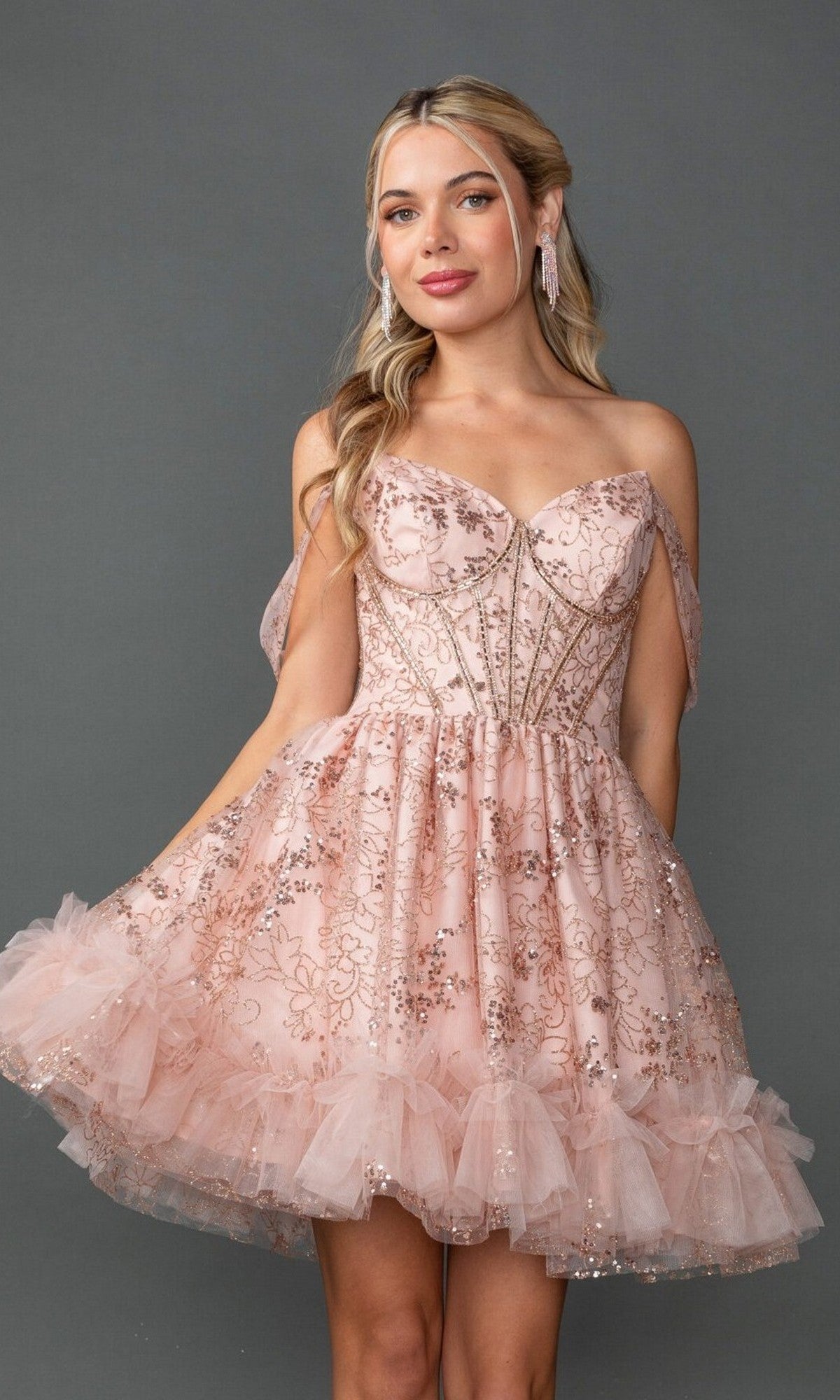 A woman models the Short Prom Dress: Dancing Queen 3422—a strapless blush pink dress with a corset bodice, sequin floral embroidery, and tulle ruffles—while standing against a plain gray background.
